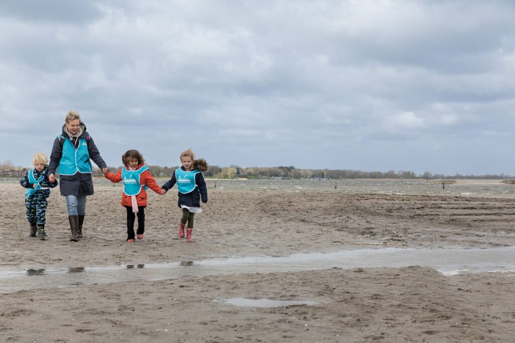 Een begeleidster loopt met drie kinderen hand in hand over het strand