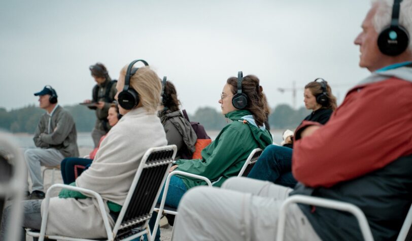 Mensen hebben een koptelefoon op en zitten in een stoel op het strand aan het IJmeer tijdens het WaterWalks evenement - Foto: Lidewij Meijer