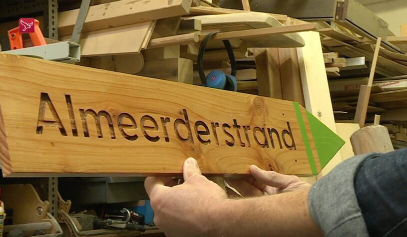 Iemand houdt een houtenbewegwijzering bordje vast met 'Almeerderstrand' erop.